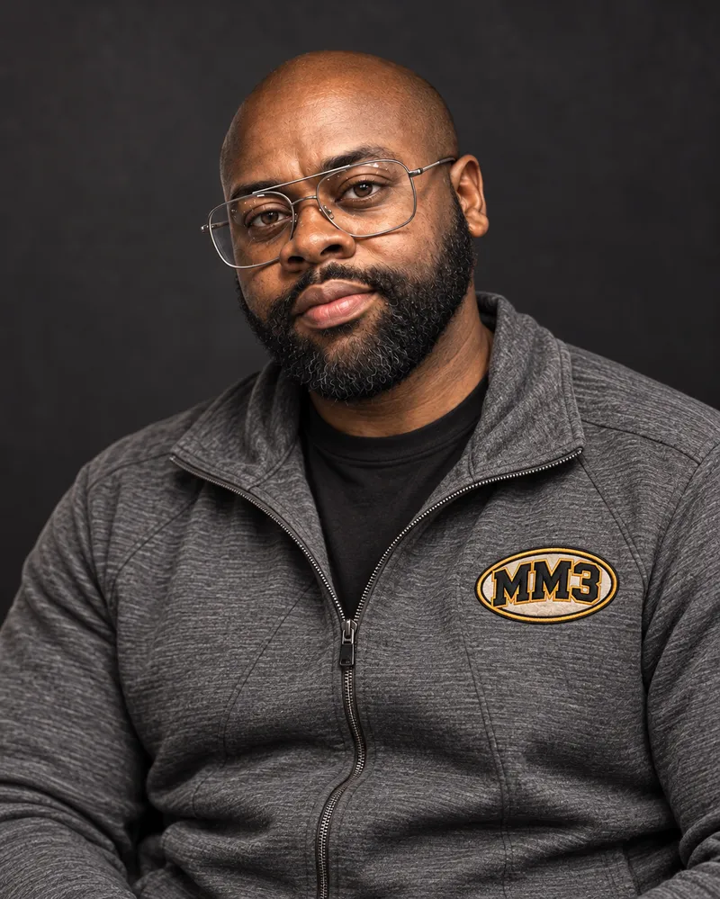 MM3 wearing glasses and a charcoal jacket with the black and gold MM3 emblem